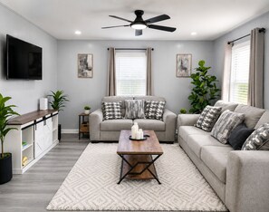 A living room with a sofa, coffee table, TV, ceiling fan, and artwork on the walls. - Modern Halethorpe Home. Sleeps 8. 4 Beds. Near BWI, UMBC & Baltimore (halethrope)