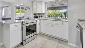 Une cuisine avec des meubles blancs, un four encastré, un évier et une fenêtre donnant sur un espace verdoyant.