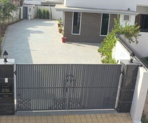 A gated entrance with a metal gate, a tiled driveway, and a building with a modern design and a small garden area. - Sant Niwas Shahpur 3BHK Villa (Mohali)