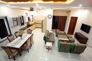 A modern living room with a dining area, green sofas, a kitchen, and a TV mounted on the wall. - Sant Niwas Shahpur 3BHK Villa (Mohali)