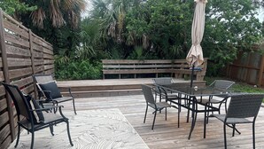 Una terraza de madera con una mesa y sillas de vidrio, rodeada por una valla de madera y abundante vegetación.