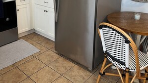 Una cocina con armarios blancos, un refrigerador de acero inoxidable, una cesta de mimbre encima, una pequeña mesa con una vela y una silla de mimbre.