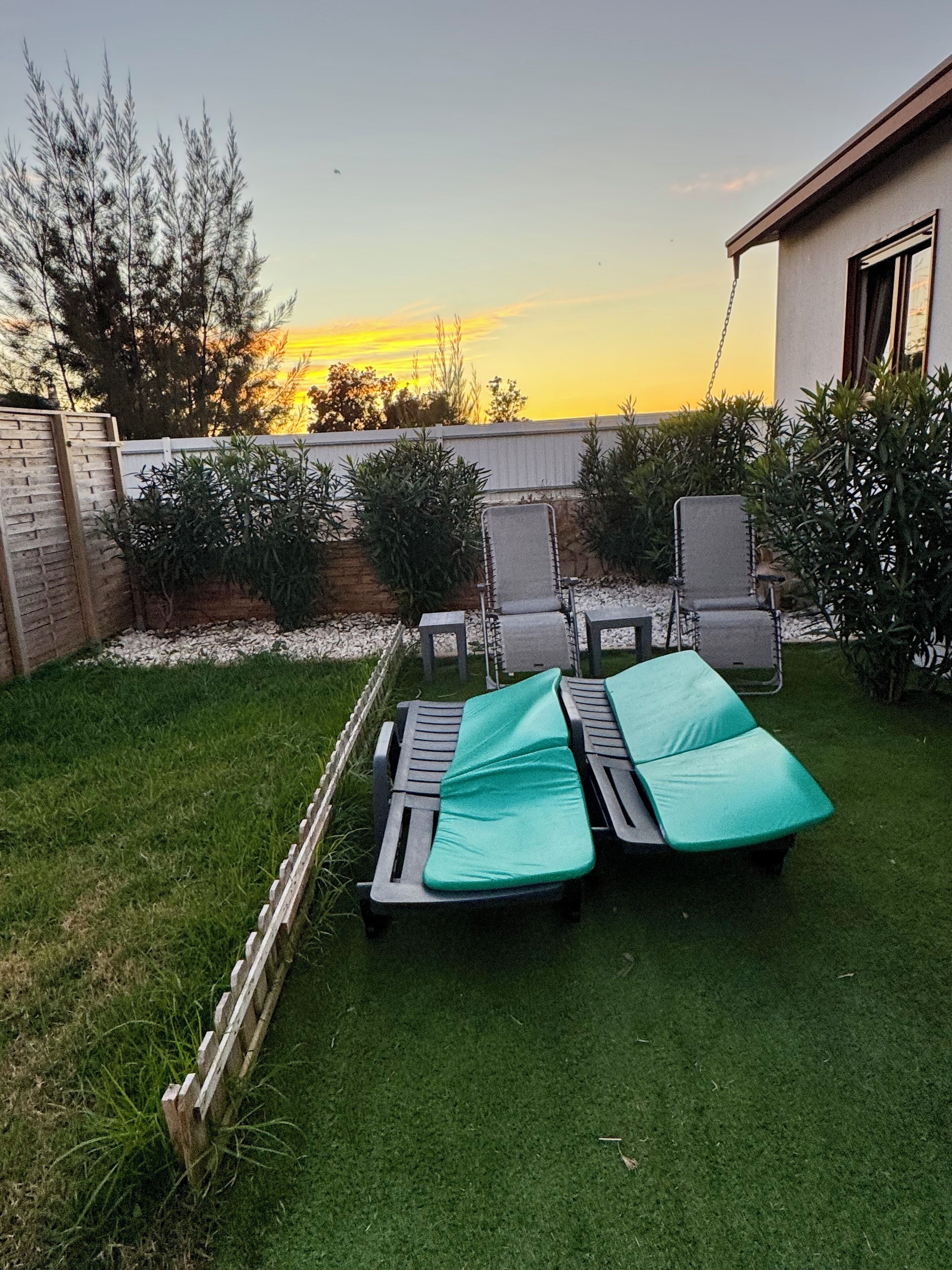 Un patio trasero con dos sillones de jardín y una mesita, rodeado por un bajo muro de ladrillo y diversas plantas.