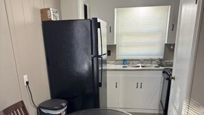 A compact kitchen with a black refrigerator, a small trash bin, a window with blinds, and a dining table with two chairs. - Cozy 1-bedroom home in the heart of Jonesboro! (Jonesboro)