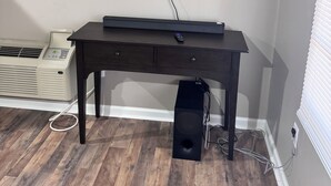 A room with a mounted flat-screen TV, a dark wooden table with a remote, a white air conditioner, and a black speaker on the floor. - Cozy 1-bedroom home in the heart of Jonesboro! (Jonesboro)