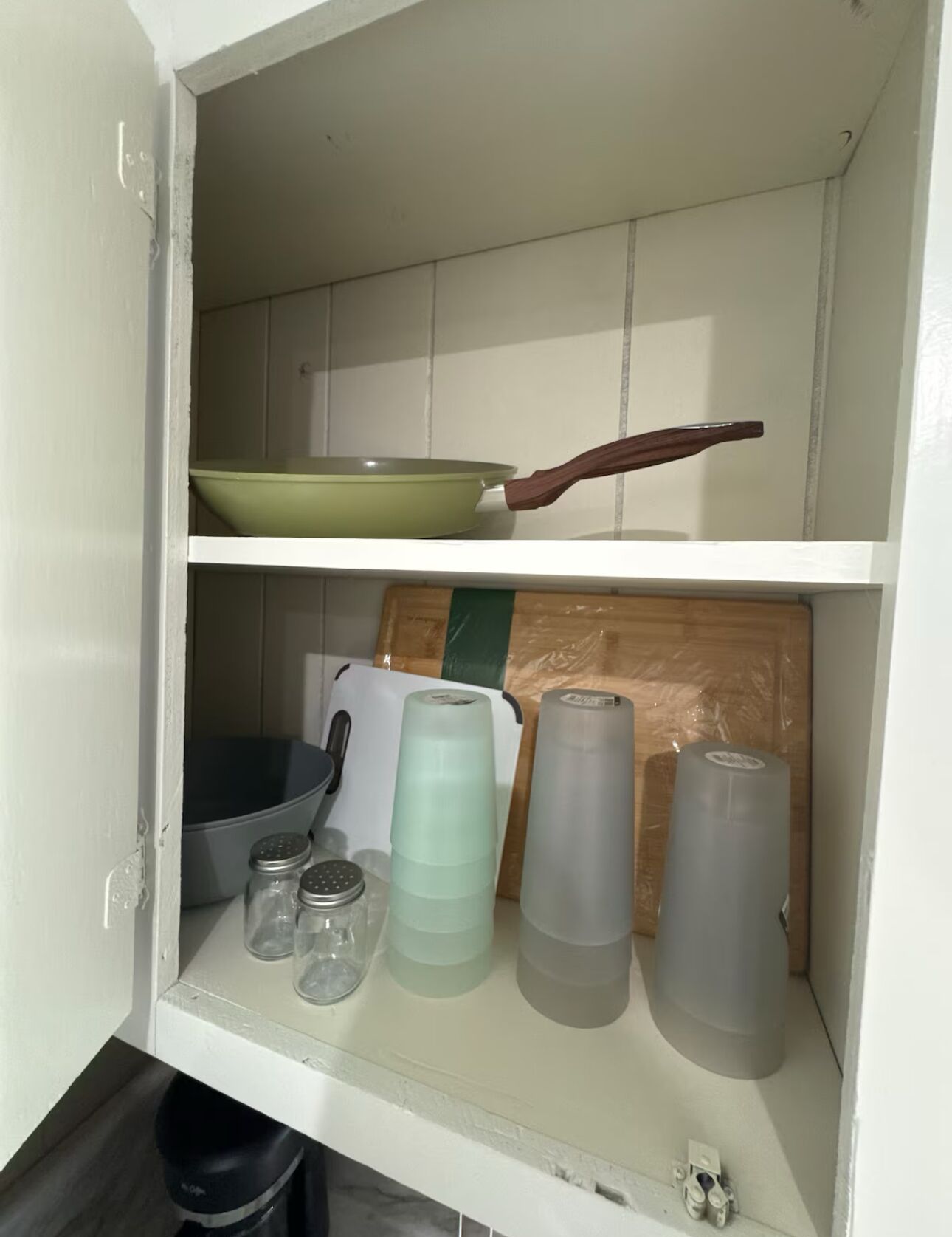 A kitchen cabinet with a green bowl, a frying pan, salt shakers, and plastic containers.