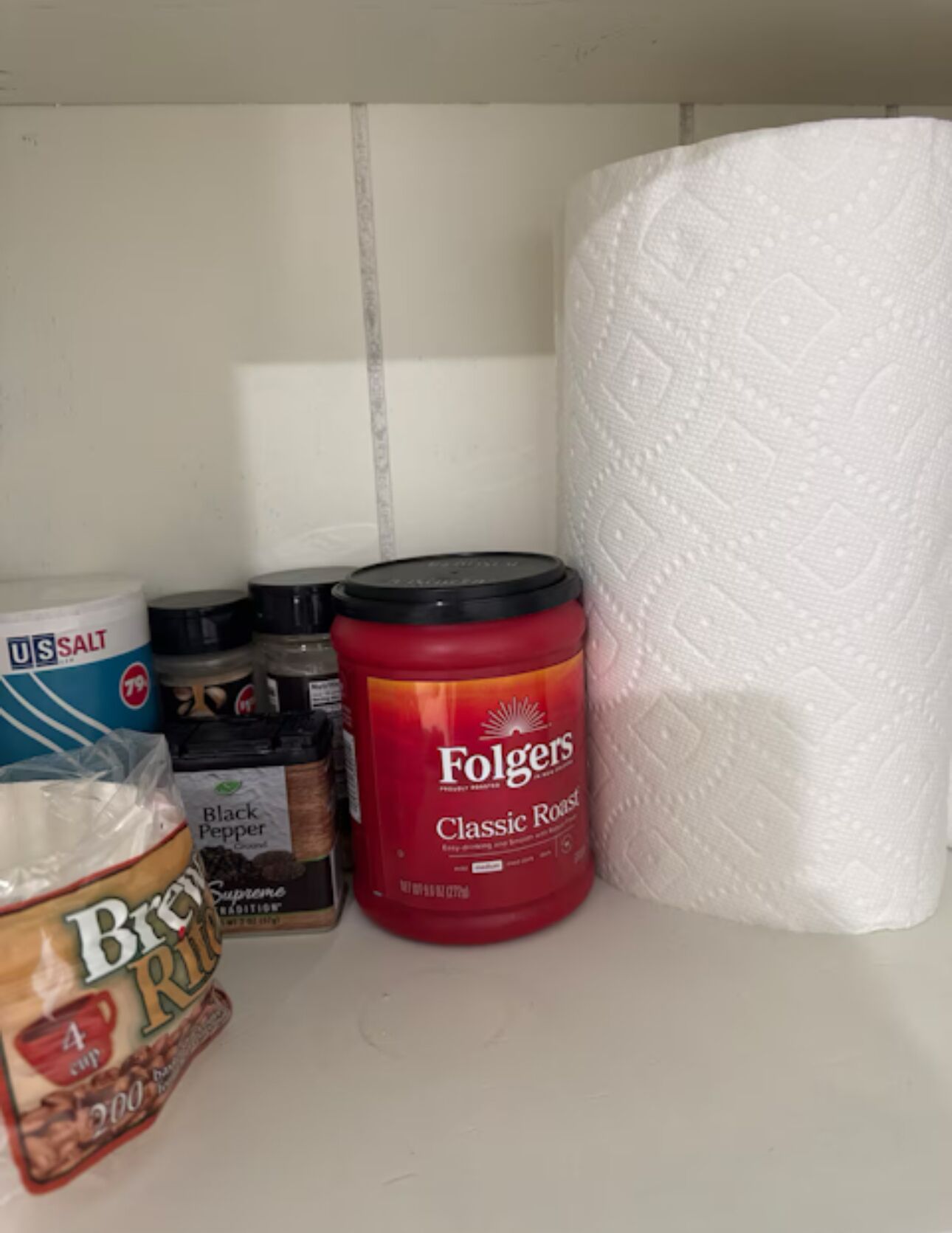 Een plank met diverse spullen, waaronder een rode Folgers koffiebus, een zak Brew Rich koffie en een pak keukenpapier.
