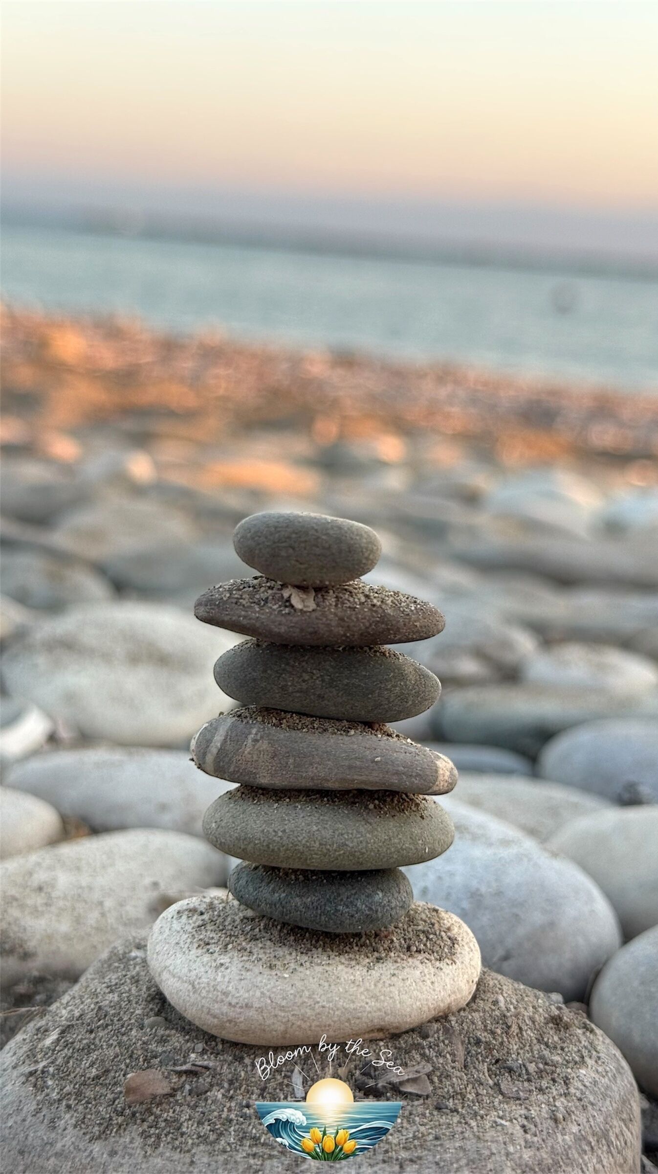 Una pila de piedras lisas y redondeadas sobre roca, con un mar tranquilo y una puesta de sol al fondo.