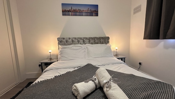 En snyggt bäddad säng med vita lakan och en grå filt, två sänglampor, en tavla med en stadsbild på väggen och ett fönster med mörka gardiner.