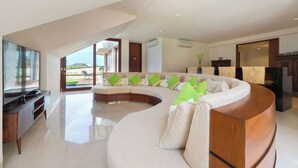 A spacious living room with a curved sectional sofa, a flat-screen TV, and a view of the outdoors. - Villa Rose by Elite Havens (The Bukit)