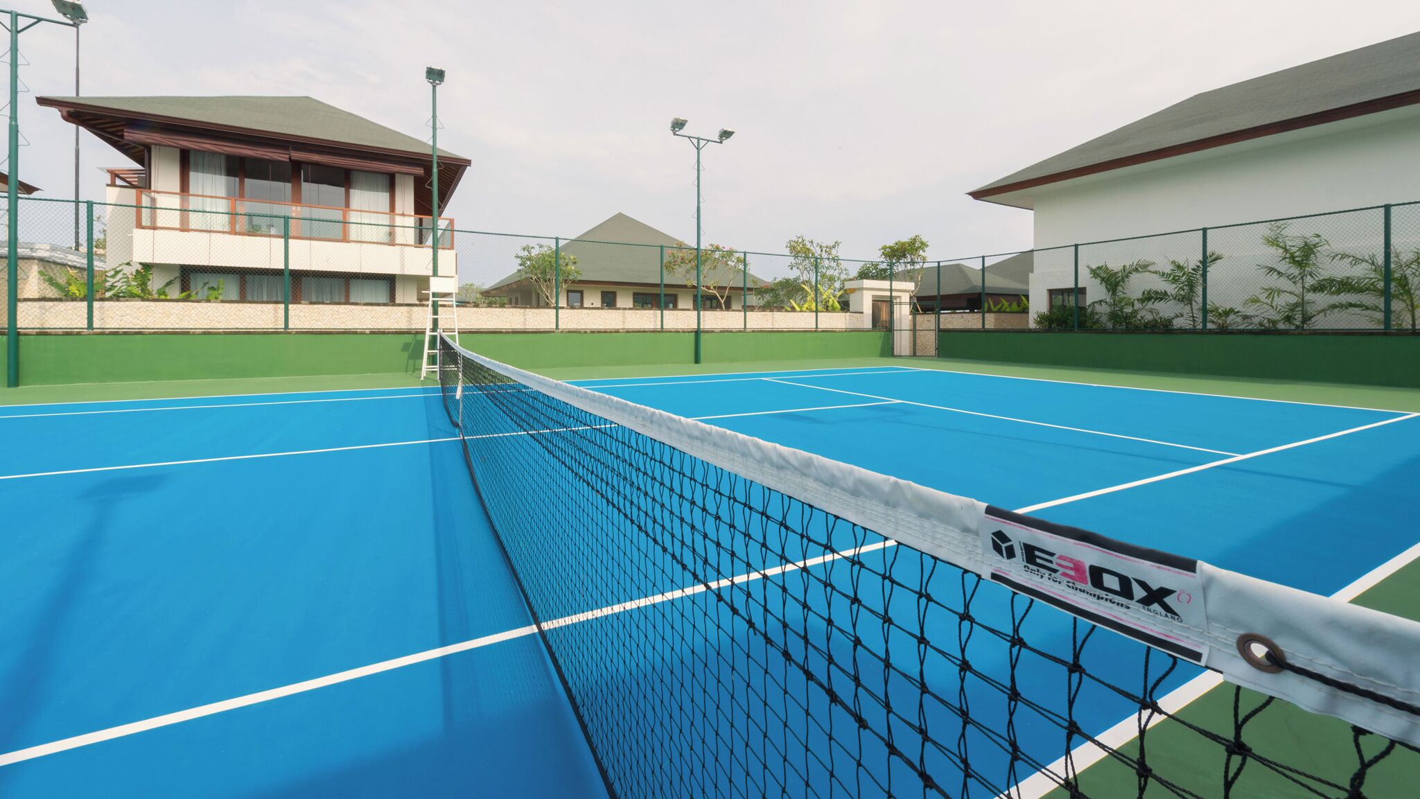 A tennis court with a net, surrounded by buildings and greenery.