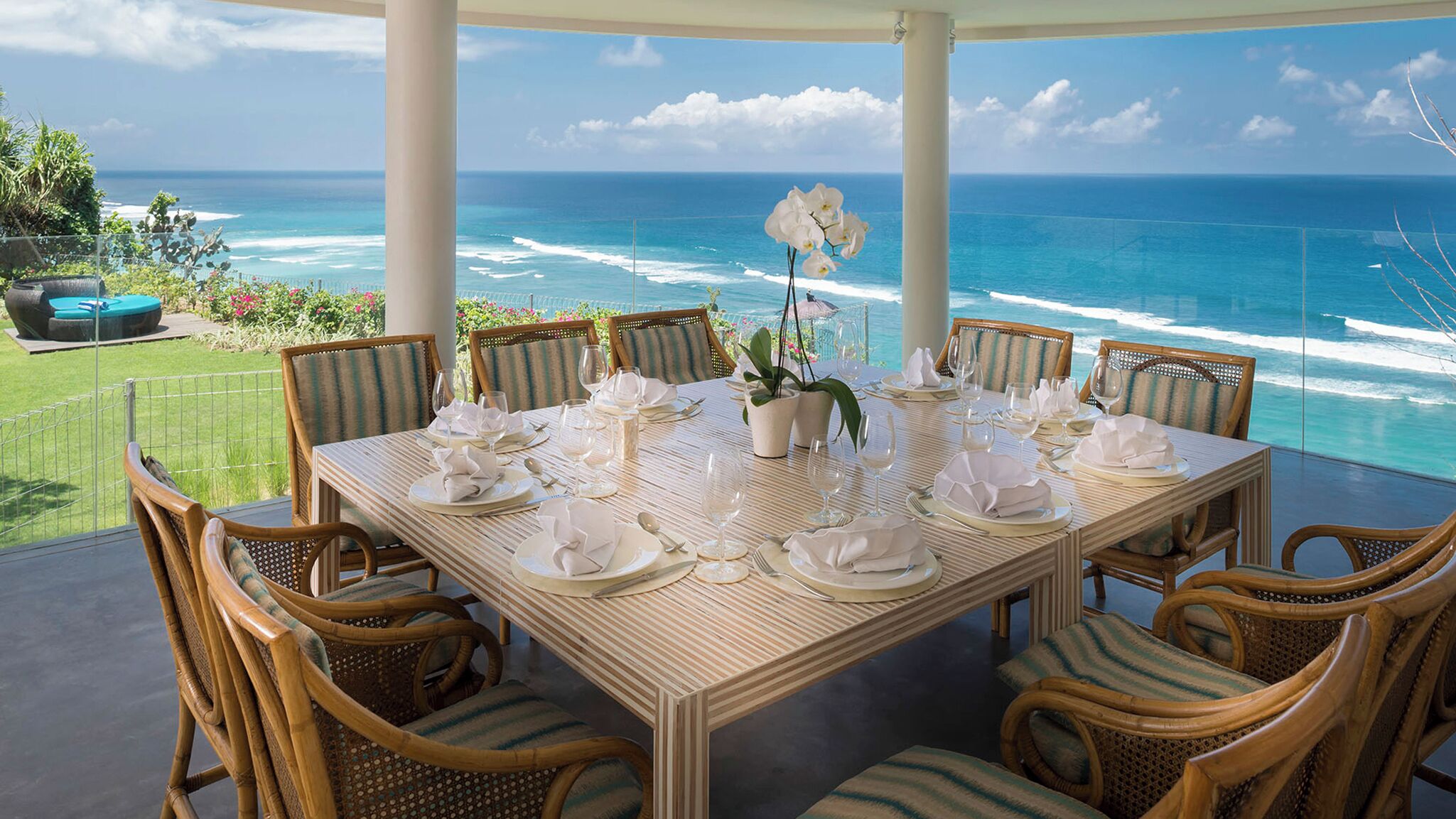 A dining table set for six with wicker chairs, overlooking a beach with ocean waves.