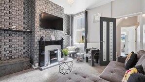 A living room with a fireplace, a flat-screen TV, a sofa, and a dining area. - 2 bed Home with Free Parking (Walsall)