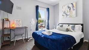 A bedroom with a bed, a desk, a chair, a TV, a window with curtains, and a painting on the wall. - 2 bed Home with Free Parking (Walsall)