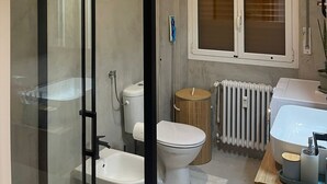 Een moderne badkamer met een glazen douchecabine, toilet, bidet, wastafel en een raam met zonwering.