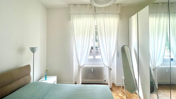 Een slaapkamer met een bed, nachtkastje, lamp, radiator en spiegel.