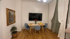 Een eethoek met een ronde tafel, vier stoelen, een kamerplant, een schilderij, een flatscreen televisie en een raam met gordijnen.