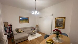 Een woonkamer met een bank, een eettafel met fruit en een vaas, een boekenkast en twee ingelijste foto's aan de muur.