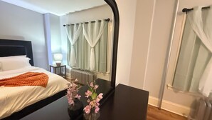 Habitación de hotel con cama, mesita de noche con lámpara, cómoda y espejo que refleja la habitación.