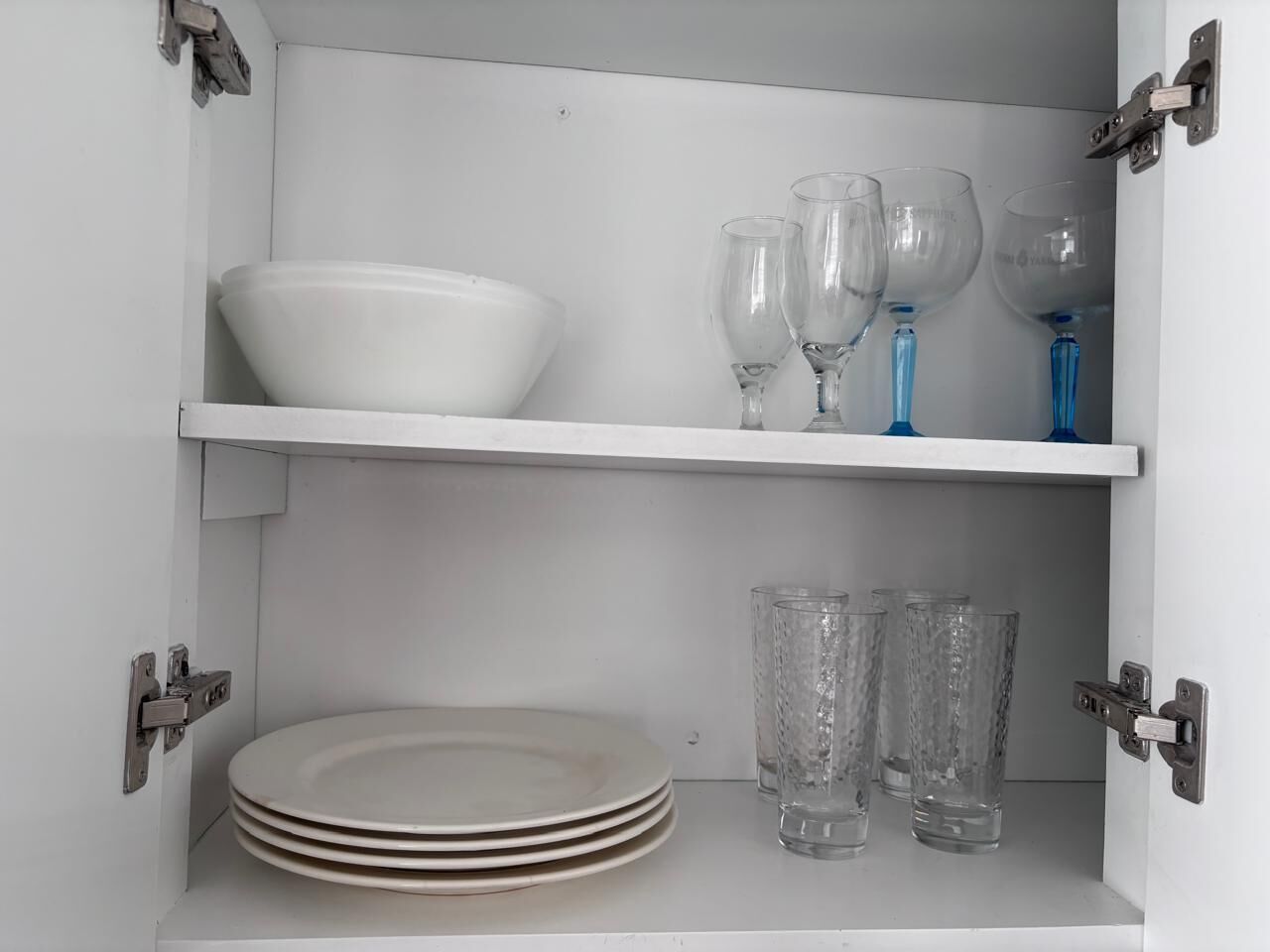 Un aparador blanco con dos estantes; el estante superior contiene un cuenco blanco y copas de vino, y el estante inferior tiene platos blancos apilados y vasos transparentes.