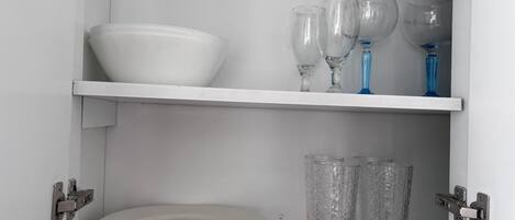 Un aparador blanco con dos estantes; el estante superior contiene un cuenco blanco y copas de vino, y el estante inferior tiene platos blancos apilados y vasos transparentes.