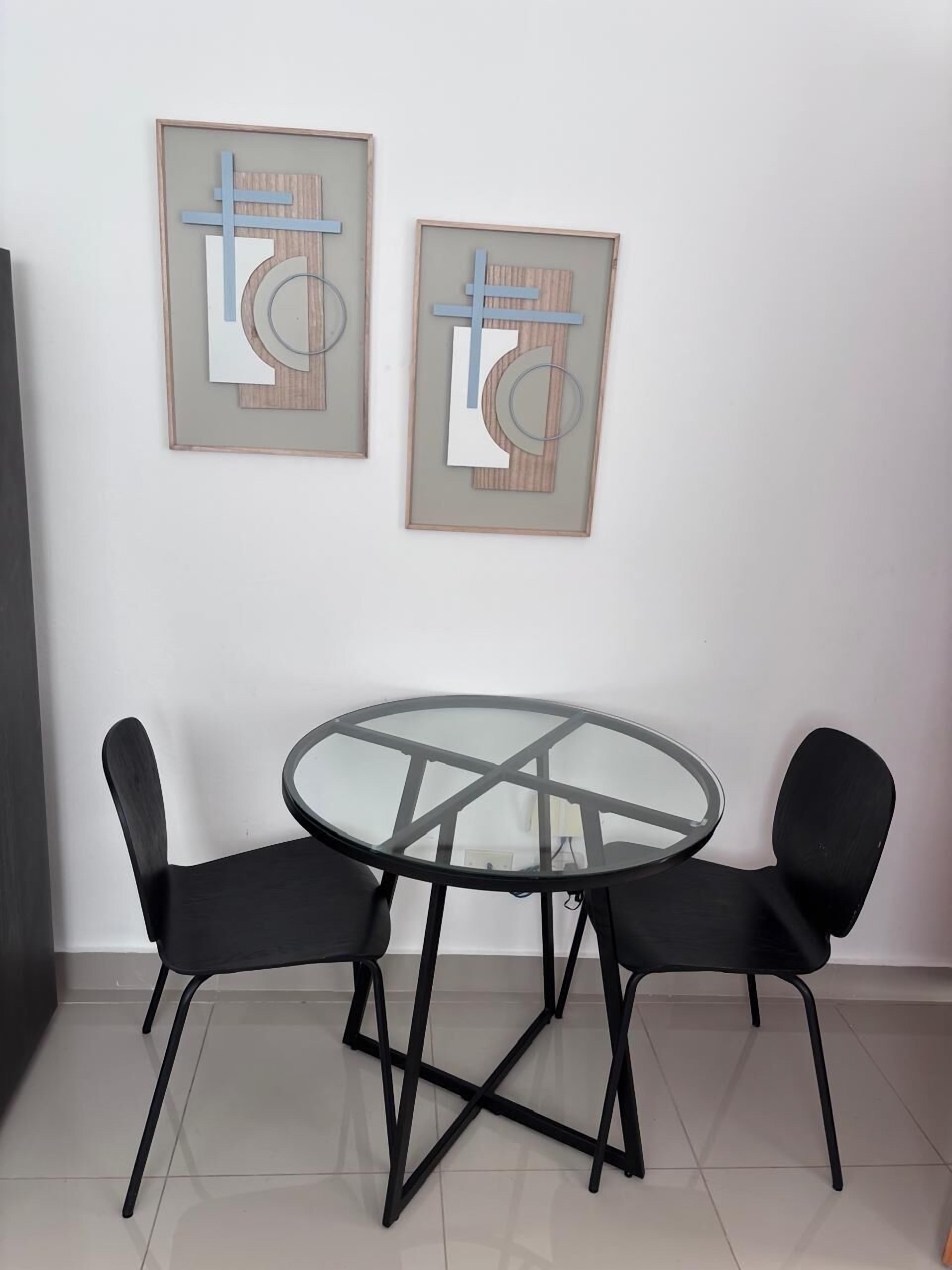 A small dining area with a round glass table and two black chairs. There are two framed abstract artworks on the wall above the table.