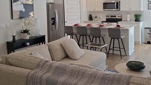 Una sala de estar moderna con un sofá beige, una mesa de centro y una cocina al fondo con armarios blancos, un refrigerador y un microondas.