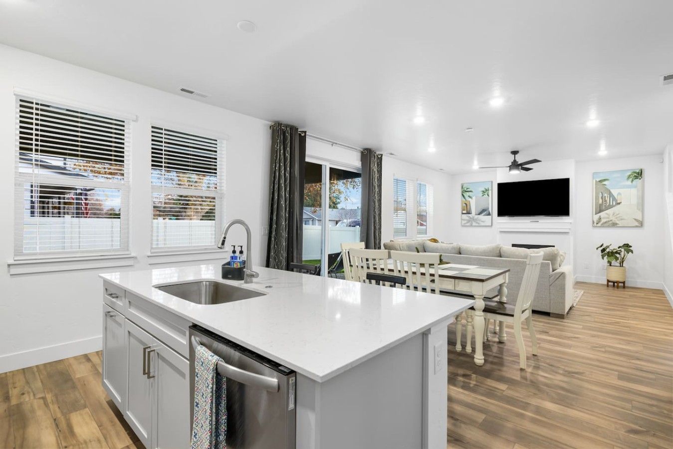 A modern kitchen with a central island, a dining area with a white table and chairs, and a living room with a sofa and TV.