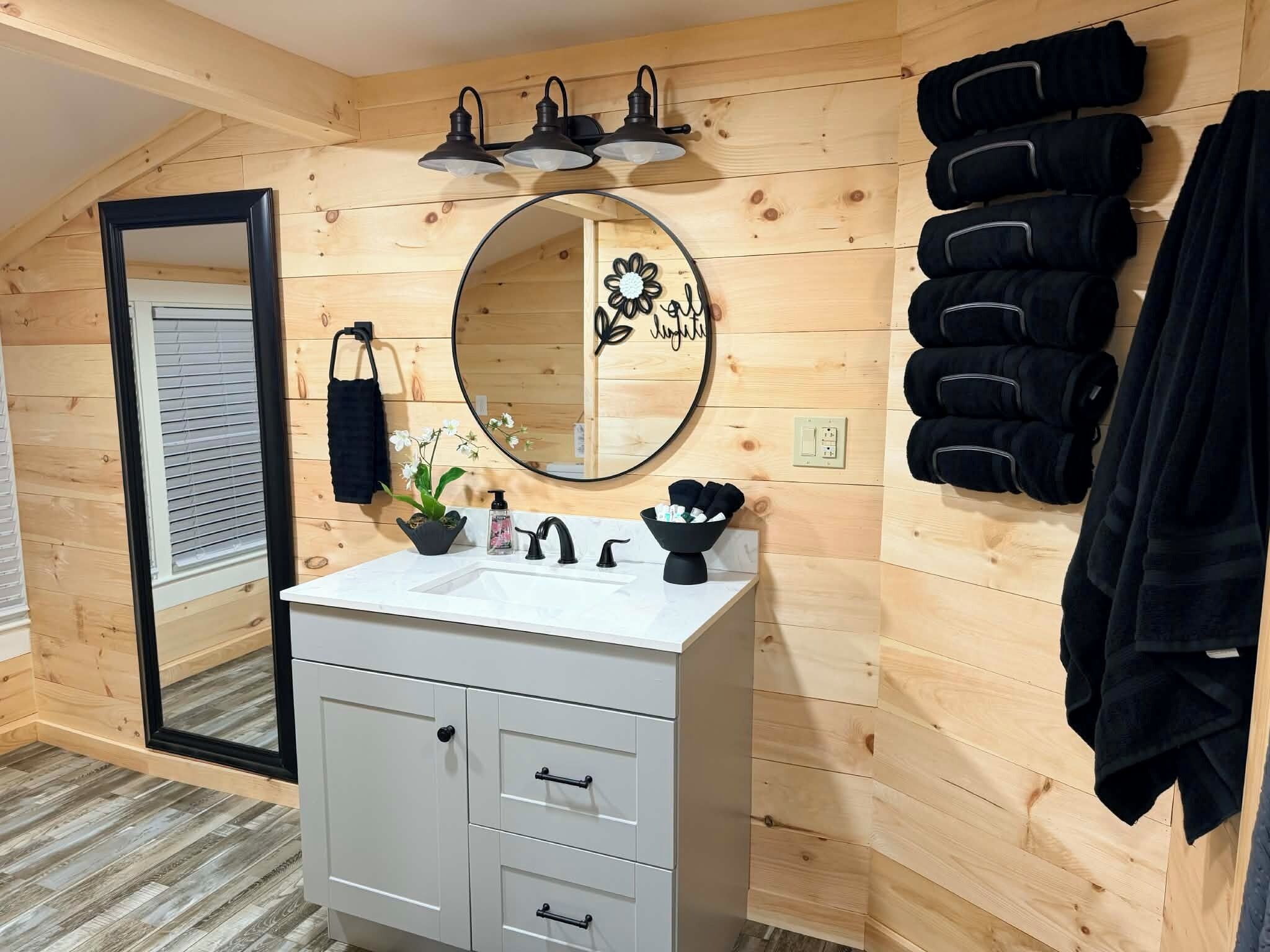 Une salle de bain aux murs lambrissés, avec un meuble-lavabo blanc, un miroir rond orné de motifs floraux et un porte-serviettes sur lequel sont disposées des serviettes noires.
