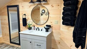 Une salle de bain aux murs lambrissés, avec un meuble-lavabo blanc, un miroir rond orné de motifs floraux et un porte-serviettes sur lequel sont disposées des serviettes noires.
