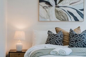 Un lit bien fait, agrémenté d’oreillers à motifs, une lampe de chevet et un grand tableau abstrait accroché au mur.
