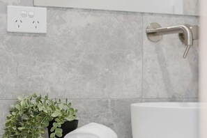Un baño con baldosas grises, un lavamanos blanco, una planta en maceta y una toalla.