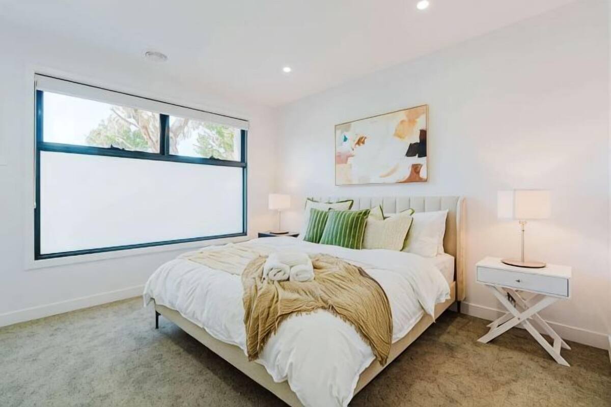 Un dormitorio con un ventanal grande, una cama con ropa de cama blanca, una mesita de noche con lámpara y un cuadro en la pared.