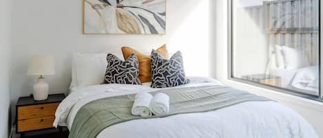 A neatly made bed with a green blanket, a pillow with a leaf pattern, and a pillow with a geometric design. There is a wall-mounted artwork above the bed and a window with a view of a balcony.