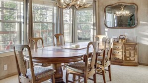 Uma sala de jantar com uma mesa redonda de madeira, seis cadeiras, um candelabro, janelas amplas e um espelho na parede.