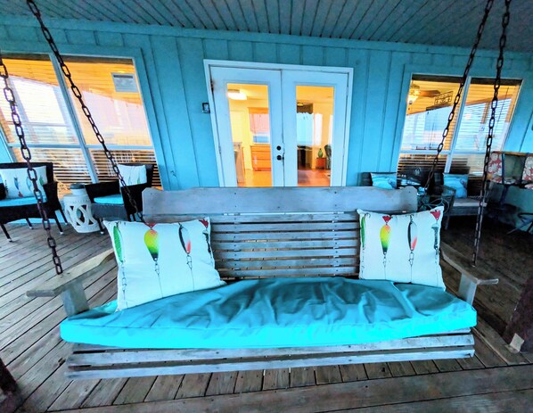 Een houten hangstoel met witte kussens, een houten terras en uitzicht op het interieur door een deur.