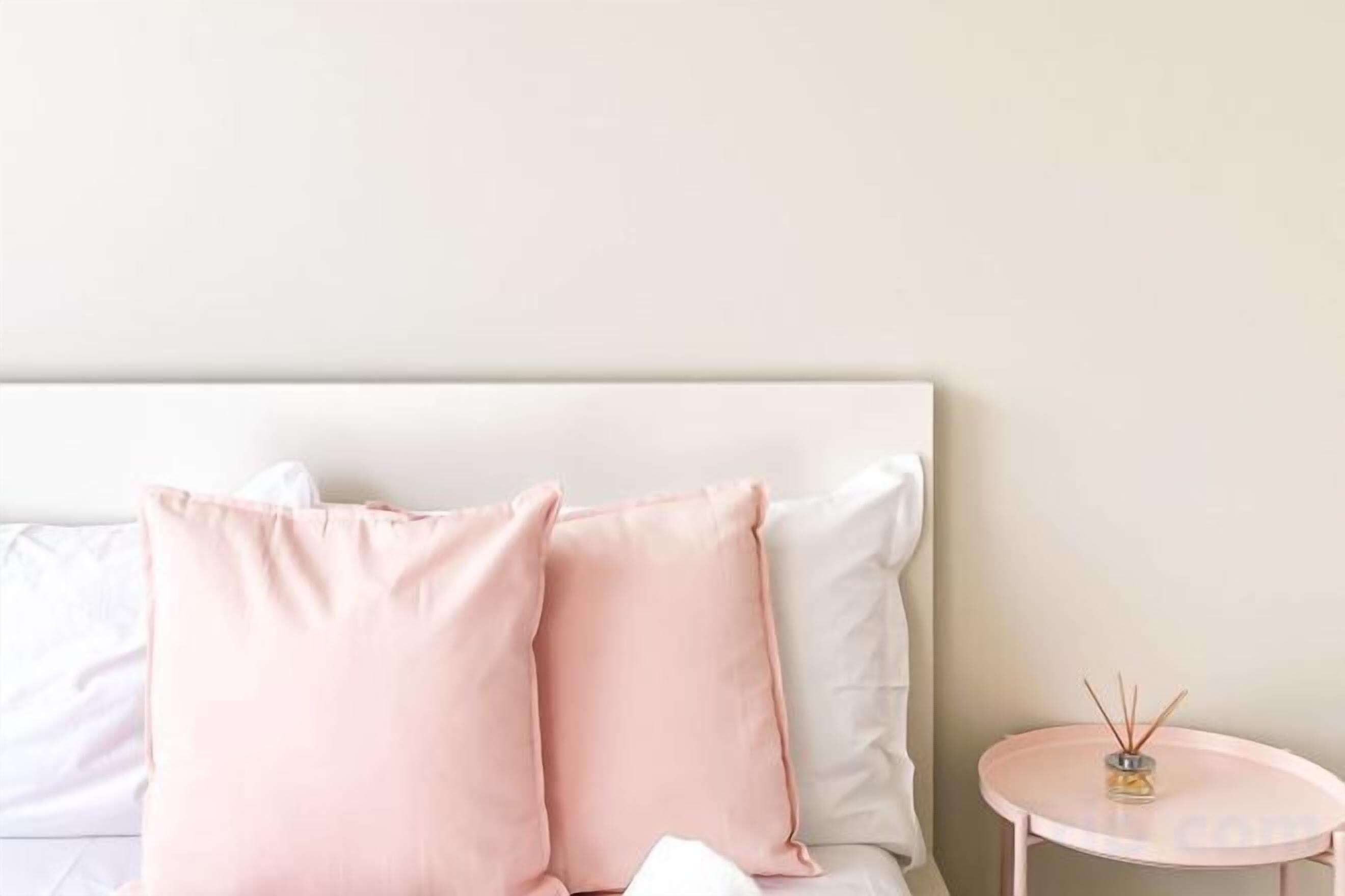 A bed with white and pink pillows, a small round table with a candle, and a plain wall.