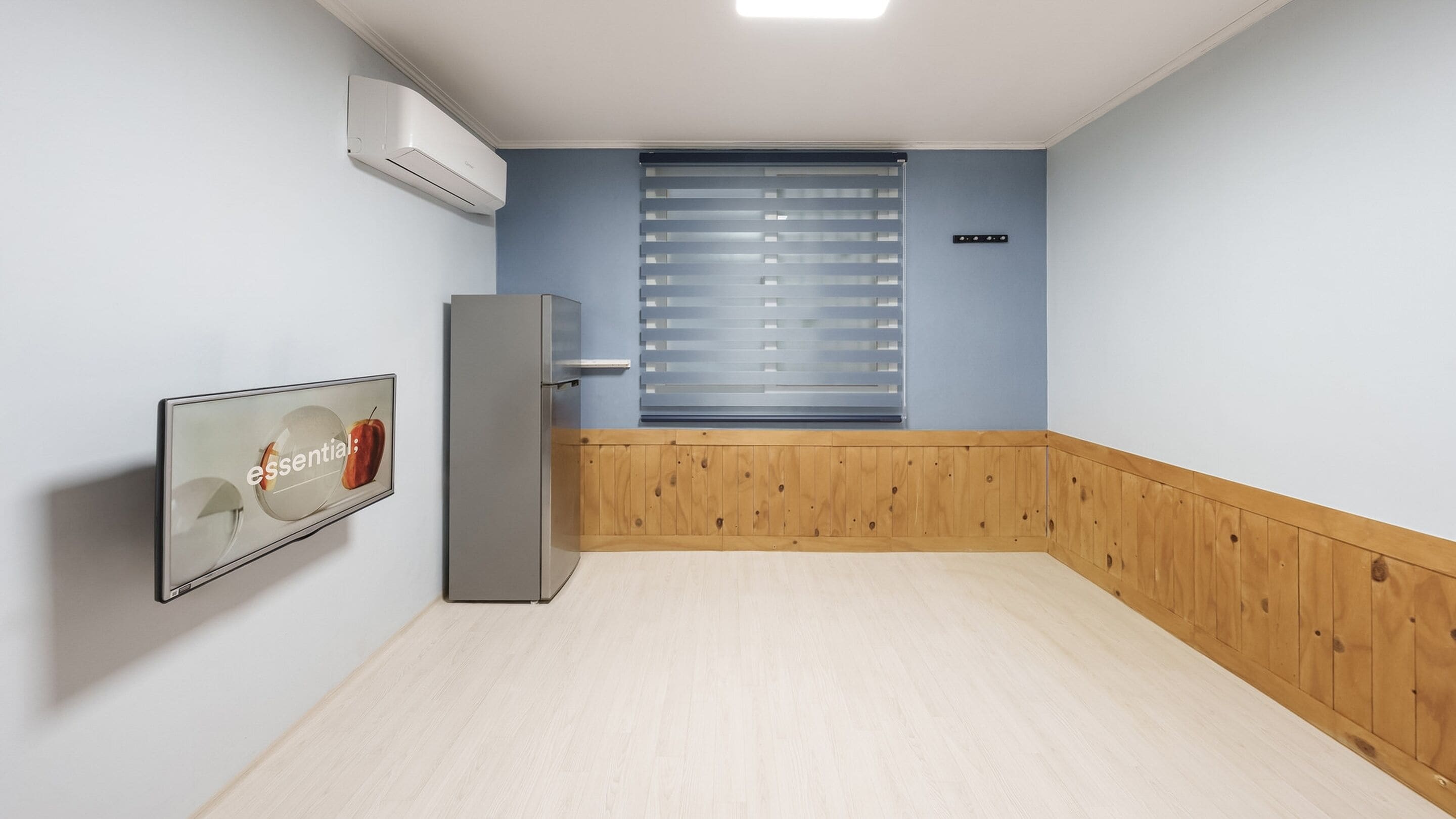 Una habitación con piso de madera, un equipo de aire acondicionado blanco, un refrigerador, una ventana con persianas y un cuadro en la pared.