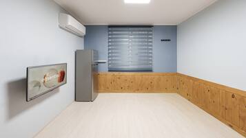 Una habitación con piso de madera, un equipo de aire acondicionado blanco, un refrigerador, una ventana con persianas y un cuadro en la pared.
