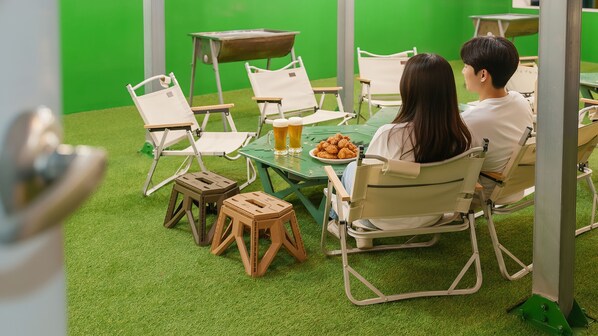 A room with green artificial grass, white folding chairs, a table with a plate of food and two beer mugs, and a green screen in the background. - Boryeong Jeil Pension (Boryeong)