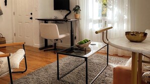 Un salon moderne avec un plancher en bois, un tapis gris, une table à manger avec un saladier, un canapé, un bureau avec une chaise et un téléviseur fixé au mur.