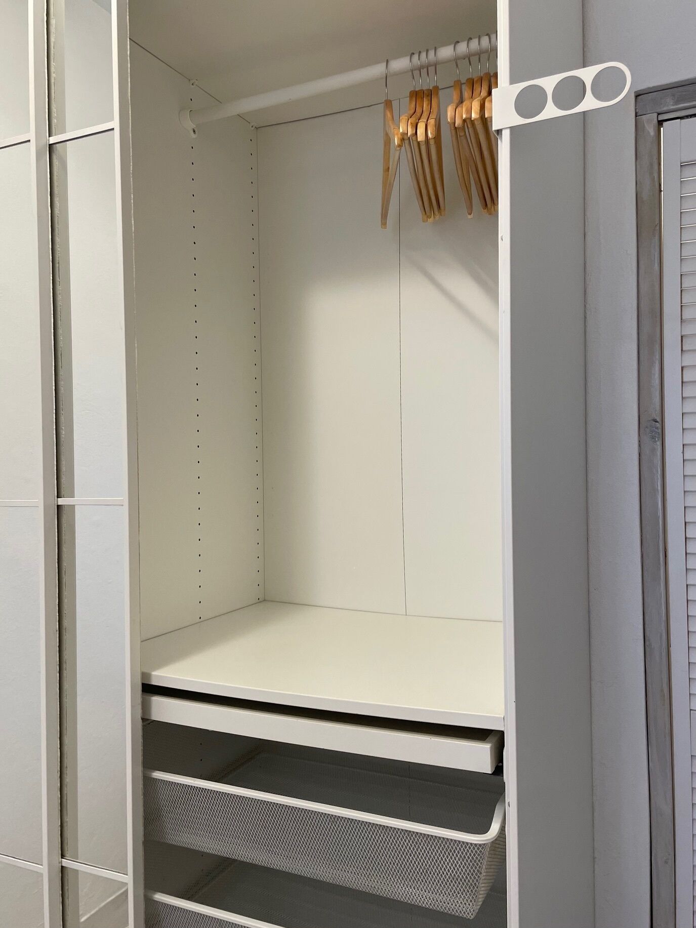 Un armario blanco con repisas y espacio para colgar, una cesta de malla metálica y perchas de madera.