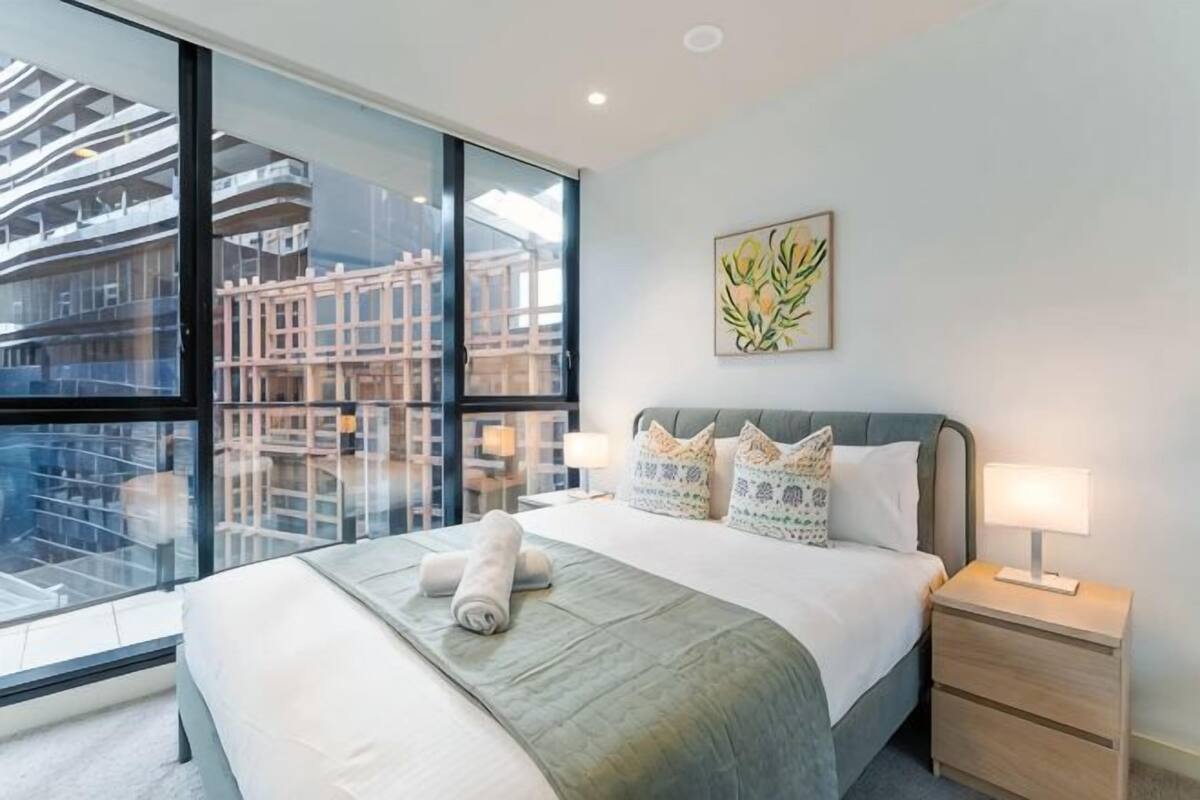 A hotel room with a large bed, a nightstand, a wall-mounted lamp, and a window with a view of buildings.