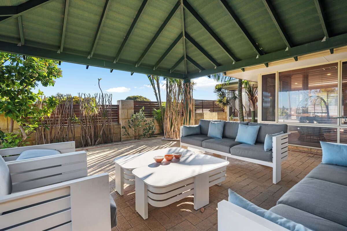 Un patio techado al aire libre con un conjunto de sofás, una mesa de centro y vistas del jardín y el edificio circundantes.