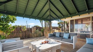 A covered outdoor patio with a sofa set, a coffee table, and a view of the surrounding garden and building. - Spacious 5-bedroom house with pool and park access  in serene Ocean Reef with AC (Ocean Reef)
