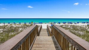 A wooden boardwalk leading to a beach with blue umbrellas and people. - Enjoy Stunning Gulf Front Cabana Club 304! Private balcony with beautiful views! (Destin)