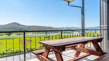 A wooden picnic table on a balcony with a view of a green field and hills.