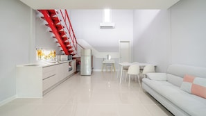 Une cuisine moderne avec un escalier rouge, un coin repas avec une table et des chaises blanches, et un canapé blanc agrémenté d’un coussin rose.