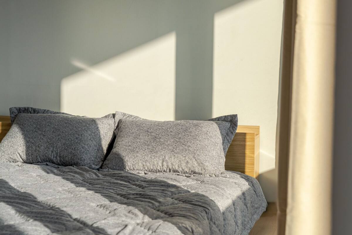 Een bed met een grijze gebreide deken en kussens, geplaatst tegen een muur met een houten hoofdbord.