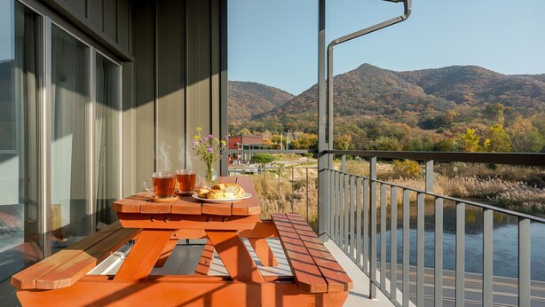 Un balcón con una mesa para dos, dos tazas de té, un plato de muffins y un jarrón de flores. El balcón tiene vistas a un río y montañas.